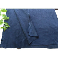 Un morceau de microfibre Longan Craft France Matière suédine au mètre, bleu foncé et extensible dans 4 directions, est posé à plat avec un coin replié. Des feuilles vertes apparaissent en haut à gauche sur un fond blanc uni.