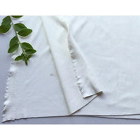 Un morceau de Matière suédine microfibre au mètre de Longan Craft France est posé à plat avec un coin plié et une tige verte feuillue en haut à gauche. Le tissu micro suédine extensible 4 directions présente des bords légèrement effilochés.