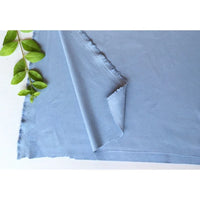 Une feuille de papier artisanal bleu clair aux bords rugueux est partiellement décollée sur une surface blanche. Plusieurs feuilles vertes et une bande de Longan Craft France Matière suédine microfibre au mètre reposent dans le coin supérieur gauche.