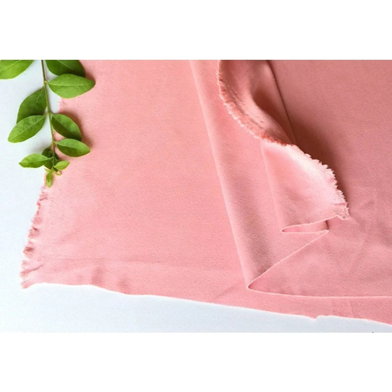 Un morceau de microfibre Matière suédine au mètre de Longan Craft France, souple et extensible dans 4 directions, est disposé avec des bords effilochés et une petite branche verte feuillue dans le coin supérieur gauche. Parfait pour les projets de bricolage de vêtements.