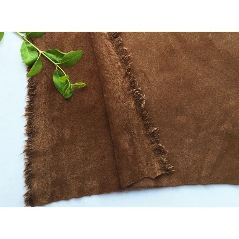 Un morceau de Matière suédine microfibre au mètre de Longan Craft France est posé à plat sur un fond clair, sa surface brune douce étant rehaussée d'une petite branche verte feuillue en haut à gauche, parfaite pour les créations vestimentaires de bricolage.