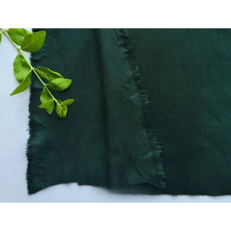 Un morceau de microfibre Matière suédine au mètre de Longan Craft France, de couleur vert foncé, est présenté à plat sur une surface blanche, avec une petite tige de plante verte et des feuilles reposant dans le coin supérieur gauche.