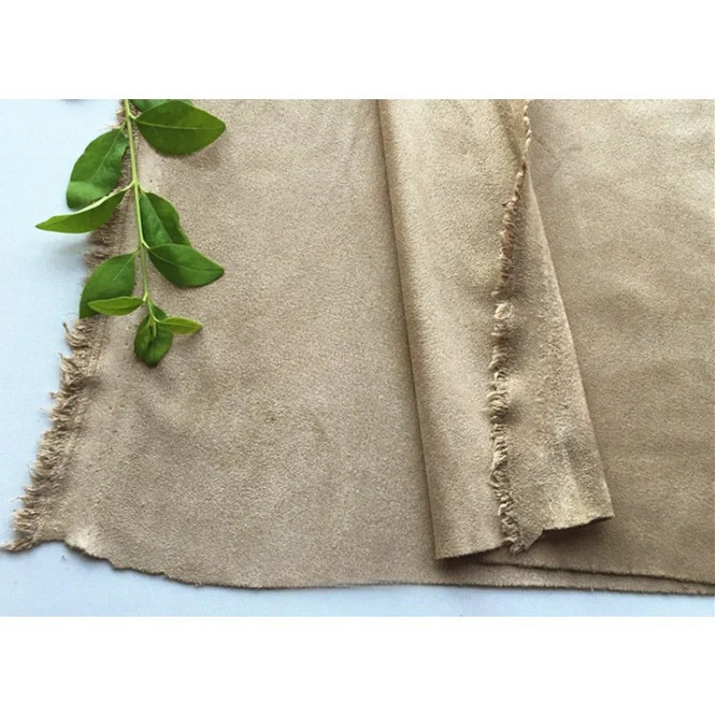Un morceau de microfibre beige Matière suédine au mètre de Longan Craft France, aux bords effilochés, est posé à plat à côté d'une petite branche verte feuillue sur une surface blanche.