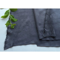 Un morceau de microfibre Longan Craft France Matière suédine au mètre, gris foncé avec des bords bruts, est posé à plat sur une surface claire. Une petite branche verte feuillue repose dans le coin supérieur gauche.