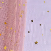 Un morceau de Tissu tulle lune et étoiles au mètre de Longan Craft France repose sur une surface blanche, avec des confettis d'étoiles dorées dispersés à droite. Ce tissu de tulle pailleté multicolore est parfait pour les costumes de scène.