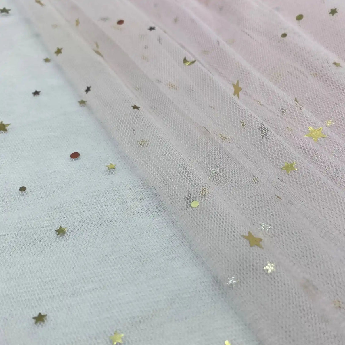Un gros plan du Tissu tulle lune et étoiles au mètre de Longan Craft France montre un tulle transparent rose pâle avec des confettis d'étoiles et de points dorés, créant un aspect délicat et scintillant - un tissu pailleté multicolore parfait pour les costumes de scène.