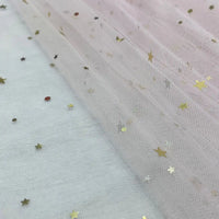 Un gros plan du Tissu tulle lune et étoiles au mètre de Longan Craft France montre un tulle transparent rose pâle avec des confettis d'étoiles et de points dorés, créant un aspect délicat et scintillant - un tissu pailleté multicolore parfait pour les costumes de scène.