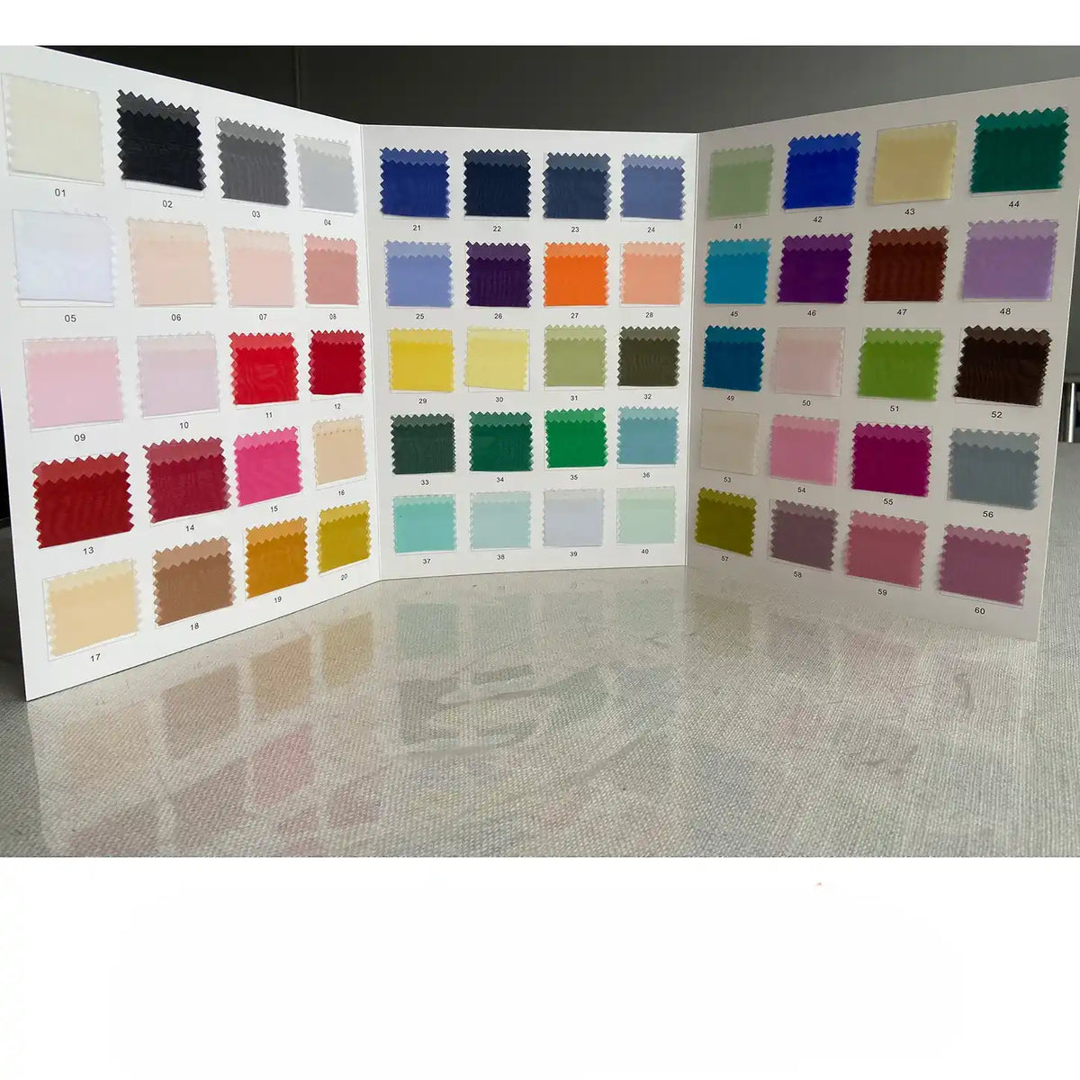 Un livre d'échantillons ouvert de Longan Craft France présente des échantillons numérotés de tissu organza multicolore en pure soie de mûrier de 8 momme, présentant une variété de nuances pour la couture de luxe sur une surface brillante.