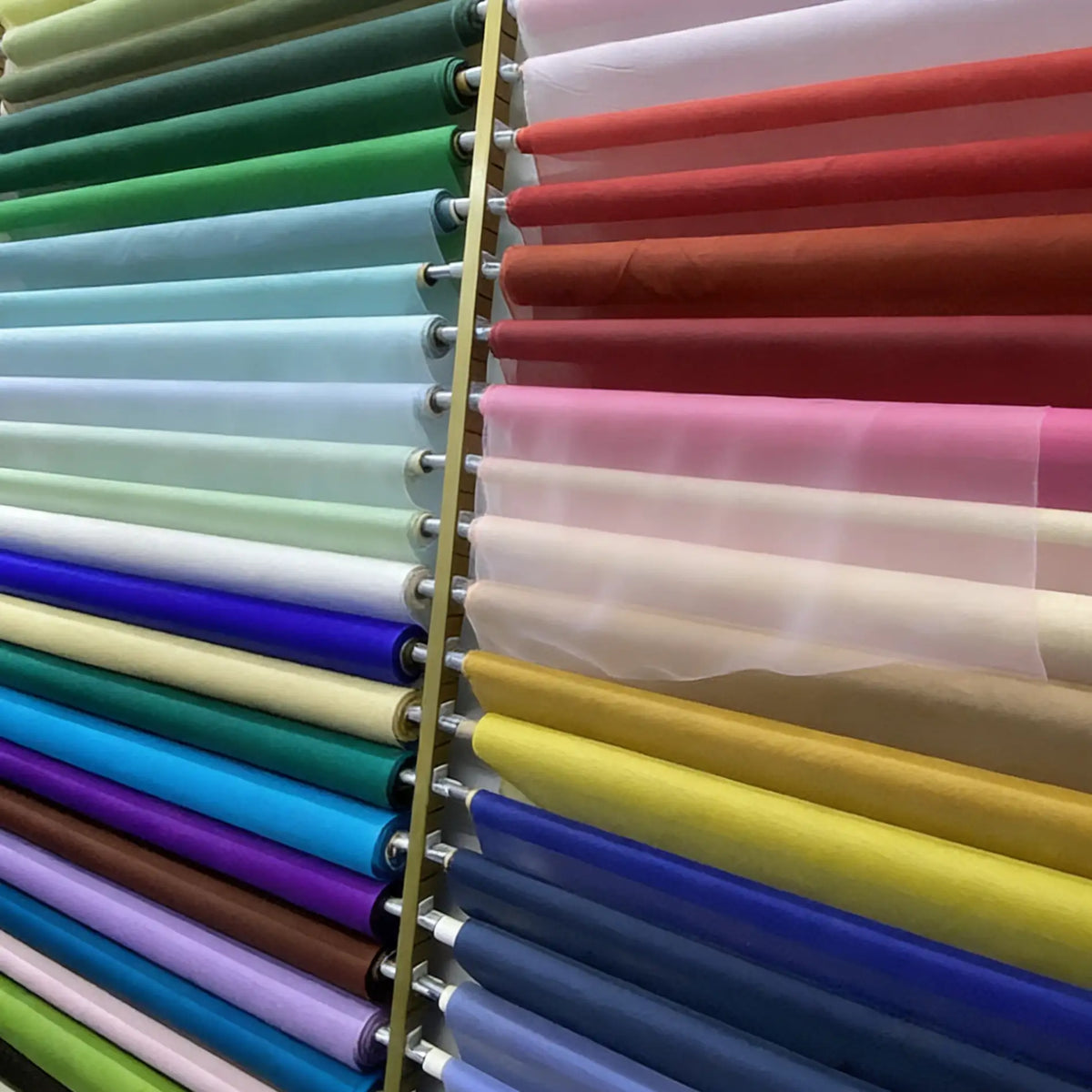 Plusieurs rouleaux d'Organza de soie multicolore 8 momme de Longan Craft France, tissu de luxe en pure soie pour robes, sont présentés verticalement sur des étagères en vert, bleu, violet, rouge, rose, jaune et blanc.