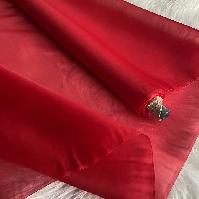Un rouleau d'Organza de soie multicolore 8 momme de Longan Craft France, pure soie de mûrier pour robes de luxe, est partiellement déroulé sur une surface texturée blanche. Le tissu léger semble translucide, avec des plis doux et une finition lisse.
