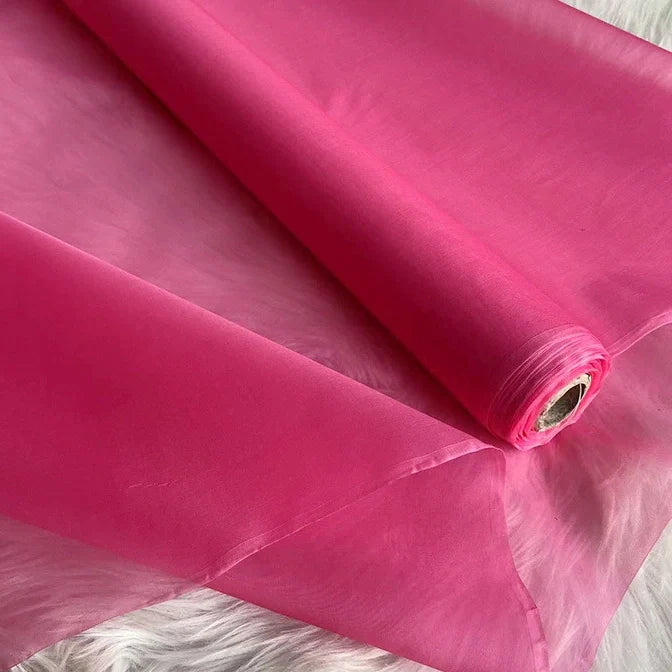 Un rouleau d'Organza de soie multicolore 8 momme de Longan Craft France - un tissu luxueux et transparent en pure soie de mûrier pour les robes - est partiellement déroulé sur une surface texturée blanche, montrant sa qualité lisse, légère et semi-transparente.