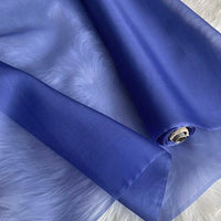 Un rouleau d'Organza de soie multicolore 8 momme de Longan Craft France, un tissu de luxe en pure soie de mûrier pour robes, est partiellement déroulé sur une surface texturée blanche. La matière transparente est légère, brillante et translucide.