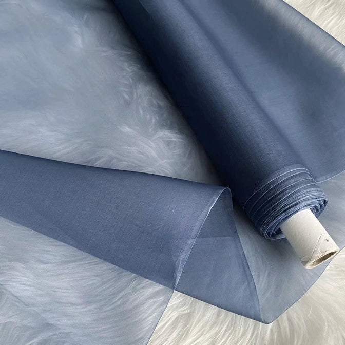Un rouleau d'Organza de soie multicolore 8 momme de Longan Craft France, fabriqué à partir de pure soie de mûrier, est partiellement déroulé sur une surface blanche et douce. Ce tissu luxueux est transparent et lisse, idéal pour les robes élégantes.
