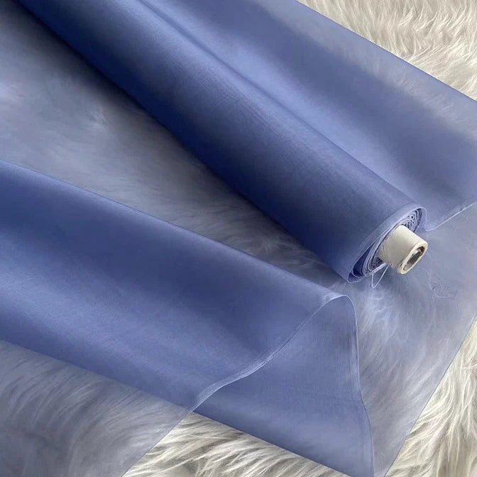 Un rouleau d'Organza de soie multicolore 8 momme de Longan Craft France est partiellement déroulé sur une surface blanche moelleuse, montrant son aspect translucide et lisse de pure soie de mûrier - un tissu de luxe parfait pour les robes.