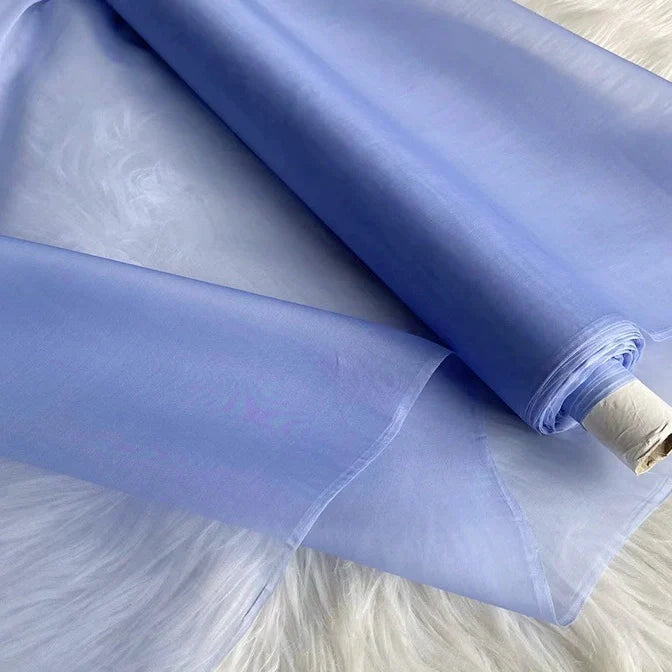 Un rouleau d'"Organza de soie multicolore 8 momme au mètre" de Longan Craft France, tissu de pure soie de mûrier pour robes de luxe, est partiellement déroulé sur une surface blanche. Le tissu de soie apparaît translucide, avec des bords lisses visibles.