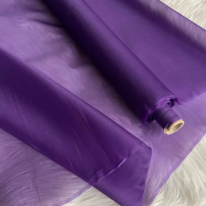 Un rouleau d'Organza de soie multicolore 8 momme de Longan Craft France, fabriqué à partir de pure soie de mûrier, est partiellement déroulé sur une surface blanche, mettant en valeur son tissu de luxe lisse et translucide, idéal pour les robes.