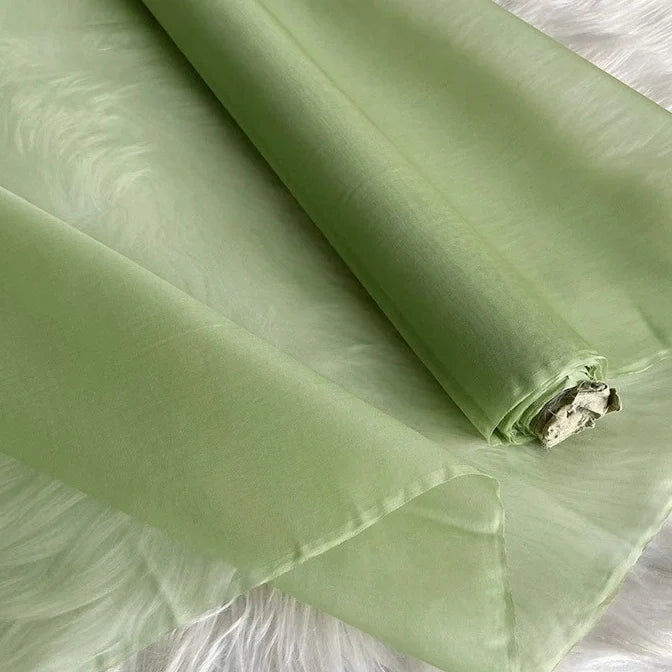 Un rouleau de Longan Craft France Organza de soie multicolore 8 momme, tissu de soie pure mûrier pour robes est partiellement déroulé sur une surface blanche douce, montrant sa matière de luxe semi-transparente vert clair.