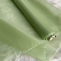 Un rouleau de Longan Craft France Organza de soie multicolore 8 momme, tissu de soie pure mûrier pour robes est partiellement déroulé sur une surface blanche douce, montrant sa matière de luxe semi-transparente vert clair.