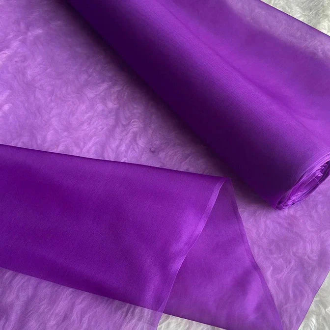 Un rouleau de Longan Craft France Organza de soie multicolore 8 momme au mètre, pure soie de mûrier, est partiellement déroulé sur une surface texturée. Ce luxueux tissu violet transparent est léger et translucide, idéal pour les peignoirs élégants.