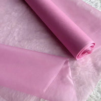 Un rouleau de Longan Craft France Organza de soie multicolore 8 momme au mètre, tissu de pure soie mûrier pour robes, est partiellement déroulé sur une surface texturée rose pâle avec les bords visibles et le tissu drapé à plat.
