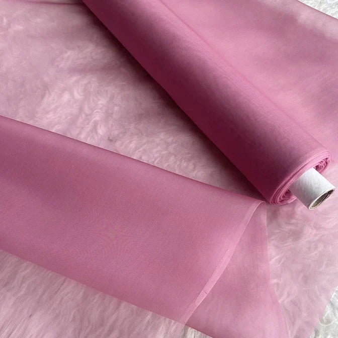 Un rouleau d'Organza de soie multicolore 8 momme au mètre de Longan Craft France, en pure soie de mûrier, est partiellement déroulé sur un duvet rose pâle. Ce luxueux tissu transparent est parfait pour confectionner des robes élégantes.