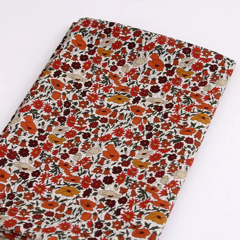 Le Longan Craft France Tissu linon de coton léger au mètre présente un imprimé floral multicolore avec des fleurs orange, rouges, crème et des feuilles vertes sur fond blanc, idéal pour la confection de vêtements d'été.