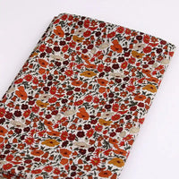 Le Longan Craft France Tissu linon de coton léger au mètre présente un imprimé floral multicolore avec des fleurs orange, rouges, crème et des feuilles vertes sur fond blanc, idéal pour la confection de vêtements d'été.
