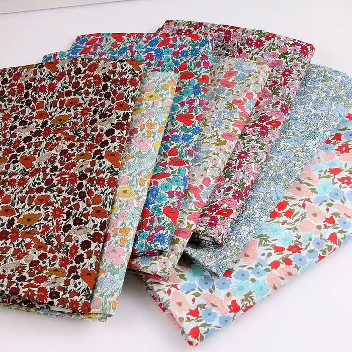Six pièces pliées du tissu linon de coton léger au mètre de Longan Craft France, aux imprimés floraux multicolores, sont disposées en éventail, idéal pour les vêtements d'été. Chaque tissu présente des motifs rouges, bleus, roses et verts sur des fonds variés.