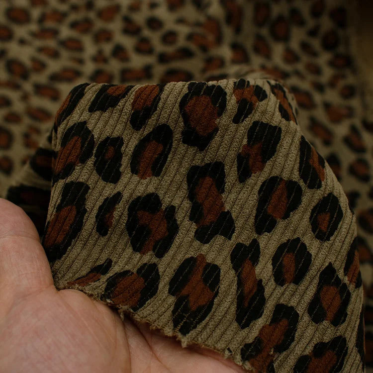 A hand holds Tissu côtelé imprimé lopard multicolore de Longan Craft France, un velours côtelé polyester doux à motif animalier, avec plus de ce tissu visible en arrière-plan.