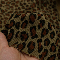 A hand holds Tissu côtelé imprimé lopard multicolore de Longan Craft France, un velours côtelé polyester doux à motif animalier, avec plus de ce tissu visible en arrière-plan.