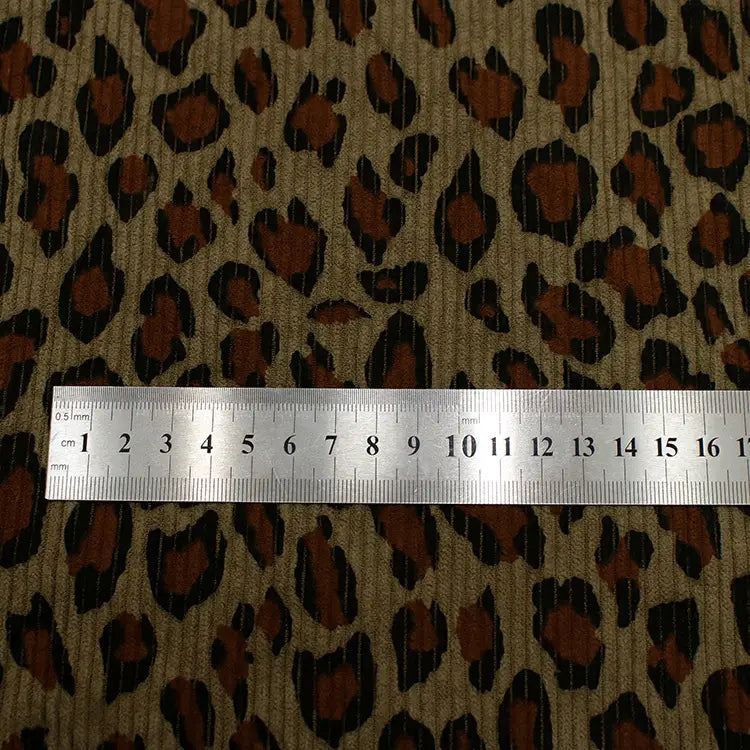Une règle métallique Longan Craft France, indiquant les centimètres 0-13, repose sur leur tissu côtelé imprimé léopard multicolore au mètre : un velours côtelé polyester avec un motif animalier doux, marron, noir et beige pour l'habillement.