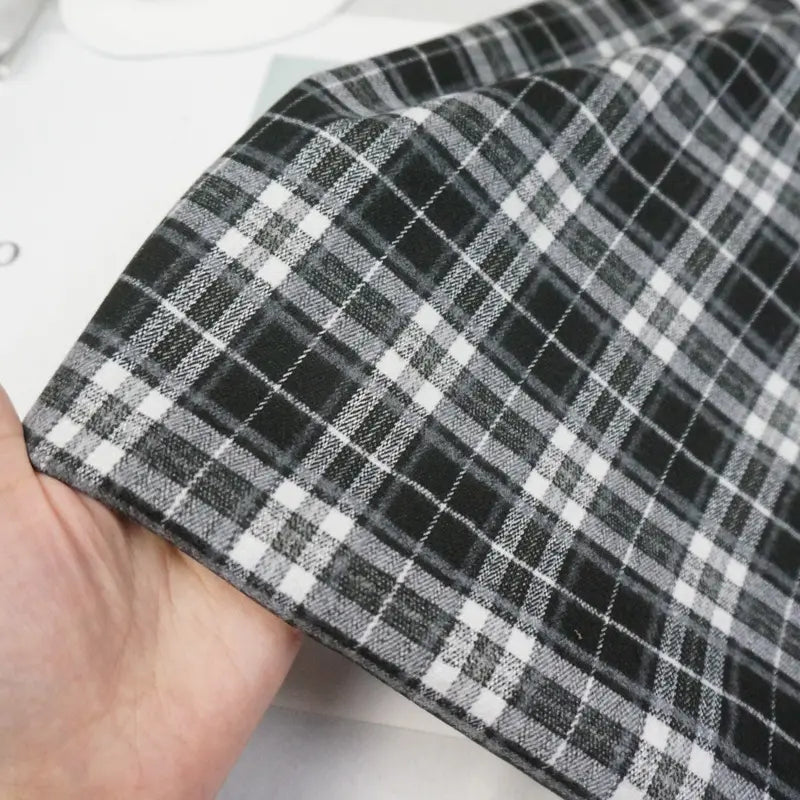 Une main tient le Tissu flanelle mammouth multicolore au mètre de Longan Craft France, un tissu flanelle à carreaux doux et brossé, idéal pour les pantalons confortables. Le motif régulièrement espacé présente des tons noirs, blancs et gris.