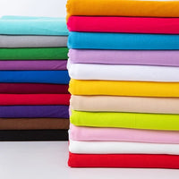 Une pile de tissus jersey polyester-élasthanne de Longan Craft France dans des couleurs telles que le rouge, le jaune, le vert, le bleu, et plus encore, présentés sur une surface blanche. Disponible au mètre pour confection de robes et projets créatifs.