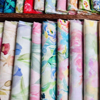 Plusieurs rouleaux de Linon de coton imprimé multicolore de Longan Craft France présentent des impressions florales pastel verticales en rose, bleu, vert et jaune doux, ajoutant un charme délicat à ce doux tissu linon de coton à fleurs.