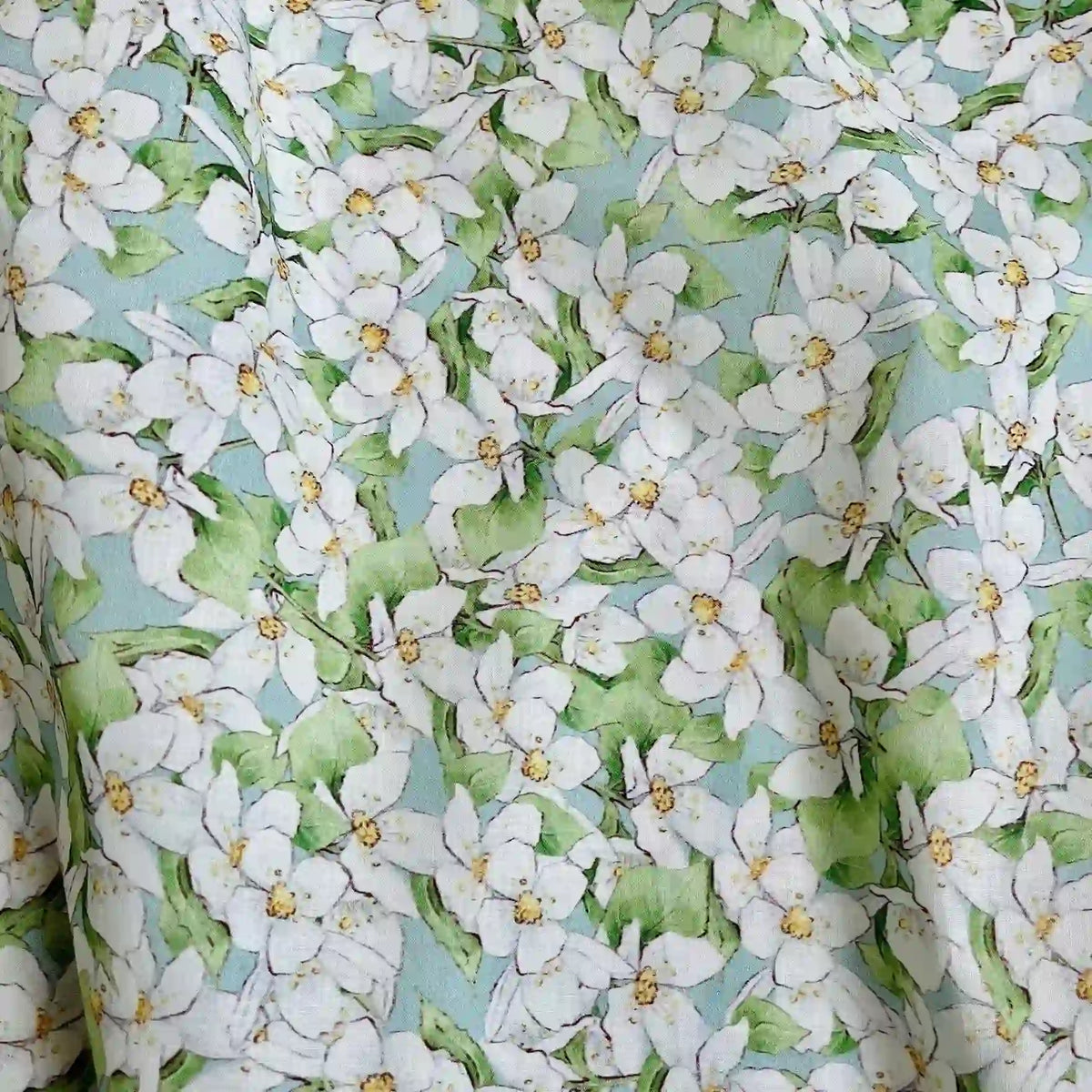 Un gros plan du Linon de coton imprimé multicolore au mètre par Longan Craft France montre des fleurs blanches avec des centres jaunes et des feuilles vertes sur un fond bleu clair, créant un charmant motif floral sur ce linon en tissu de coton doux.