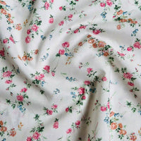 Un gros plan de Linon de coton imprimé multicolore au mètre par Longan Craft France montre un tissu de coton doux avec une impression florale dispersée - de petites fleurs roses, orange et bleues avec des tiges vertes sur le blanc, avec de légers plis et froissements.