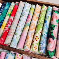Un plateau en bois contient des rouleaux de coton imprimé multicolore au mètre de Longan Craft France. Chaque rouleau de tissu présente un motif floral coloré unique dans des tons doux de rouge, de vert, de bleu, de jaune et de rose.