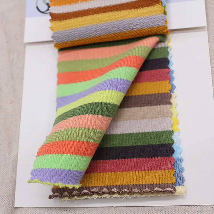 Une pile de Longan Craft France Tissu French Terry coton à rayures multicolores, avec des rayures orange, vertes, jaunes, violettes, brunes et blanches - idéales pour des sweatshirts éclatants - est présentée sur un fond blanc.