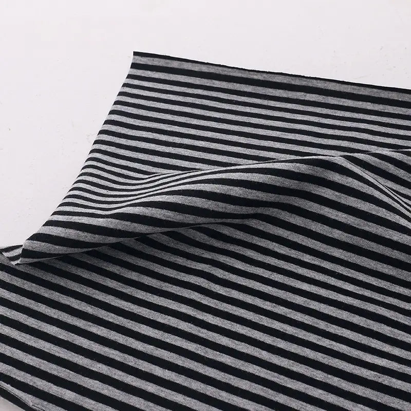 Un morceau de Tissu jersey coton rayé multicolore au mètre de Longan Craft France, jersey coton élasthanne extensible pour vêtements de sport, présente des rayures horizontales noires et gris clair et repose sur une surface blanche avec un pli.