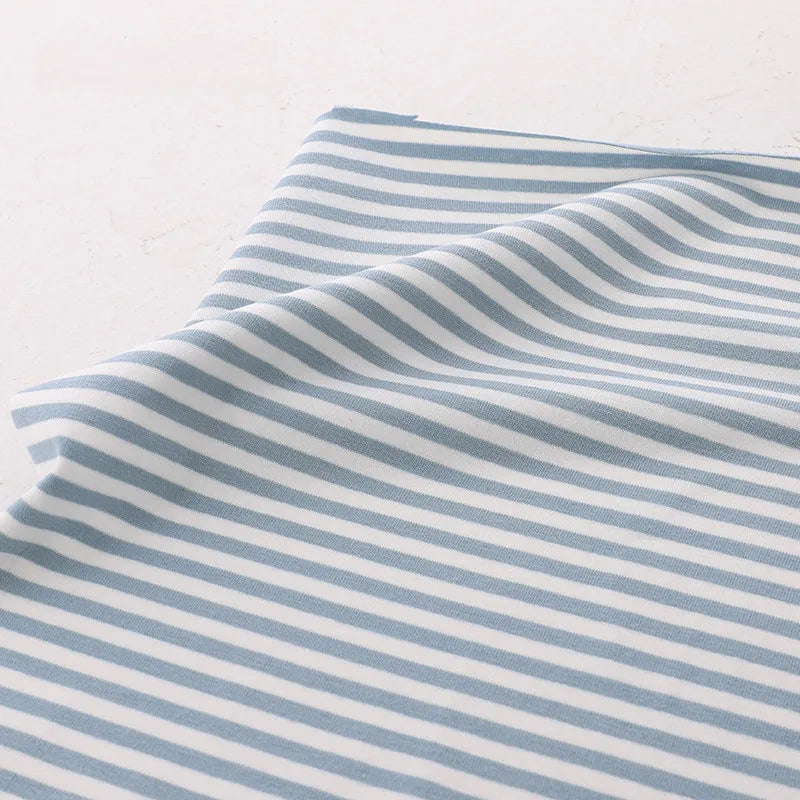 Un morceau de tissu Longan Craft France tissu jersey coton rayé multicolore au mètre, un jersey coton élasthanne doux et extensible pour vêtements de sport, avec des rayures horizontales, est posé à plat sur une surface légère avec un léger pli près d'un bord.
