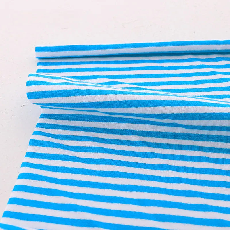 Un morceau de Tissu jersey coton rayé multicolore au mètre de Longan Craft France, avec des rayures horizontales uniformément espacées bleues et blanches, est partiellement enroulé sur une surface blanche plane, montrant la légèreté et la douceur du tissu jersey coton extensible.