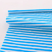 Un morceau de Tissu jersey coton rayé multicolore au mètre de Longan Craft France, avec des rayures horizontales uniformément espacées bleues et blanches, est partiellement enroulé sur une surface blanche plane, montrant la légèreté et la douceur du tissu jersey coton extensible.