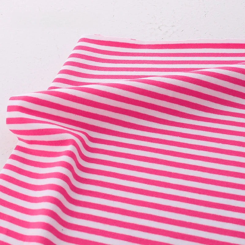Un morceau de Tissu jersey coton rayé multicolore au mètre de Longan Craft France, tissu de coton extensible pour vêtements de sport, est présenté avec des rayures horizontales roses et blanches sur une surface blanche.