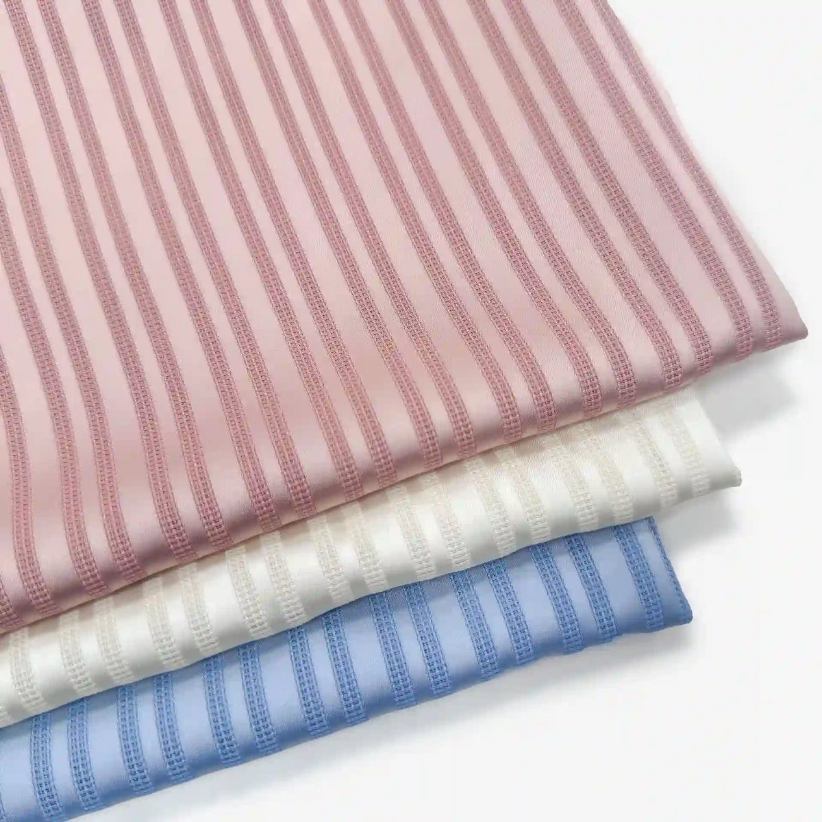 Trois tissus pliés sont empilés : un tissu rayé rose pour vêtements en haut, un tissu blanc avec des rayures similaires au milieu et, en bas, un tissu bleu rayé multicolore en polyester de Longan Craft France.