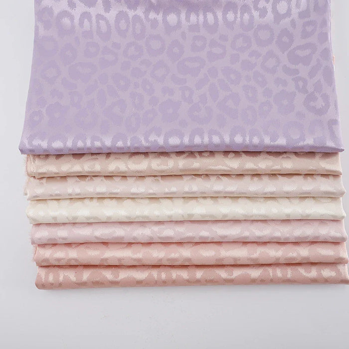 Une pile de tissu satin jacquard léopard au mètre de Longan Craft France, présentant un subtil imprimé léopard dans des tons pastel allant du violet clair au rose et au crème - un textile satiné parfait pour les chemises DIY.