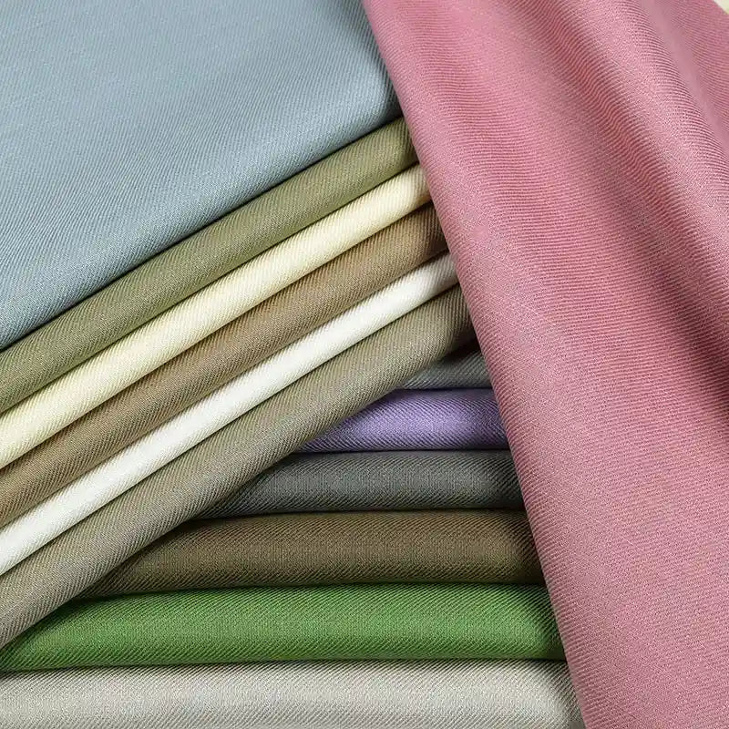Une pile de Tissu lyocell sergé coloré au mètre par Longan Craft France, 100% lyocell, présenté dans des couleurs unies superposées comme le rose, le vert, le gris, le beige et le violet - un textile confortable idéal pour les costumes.