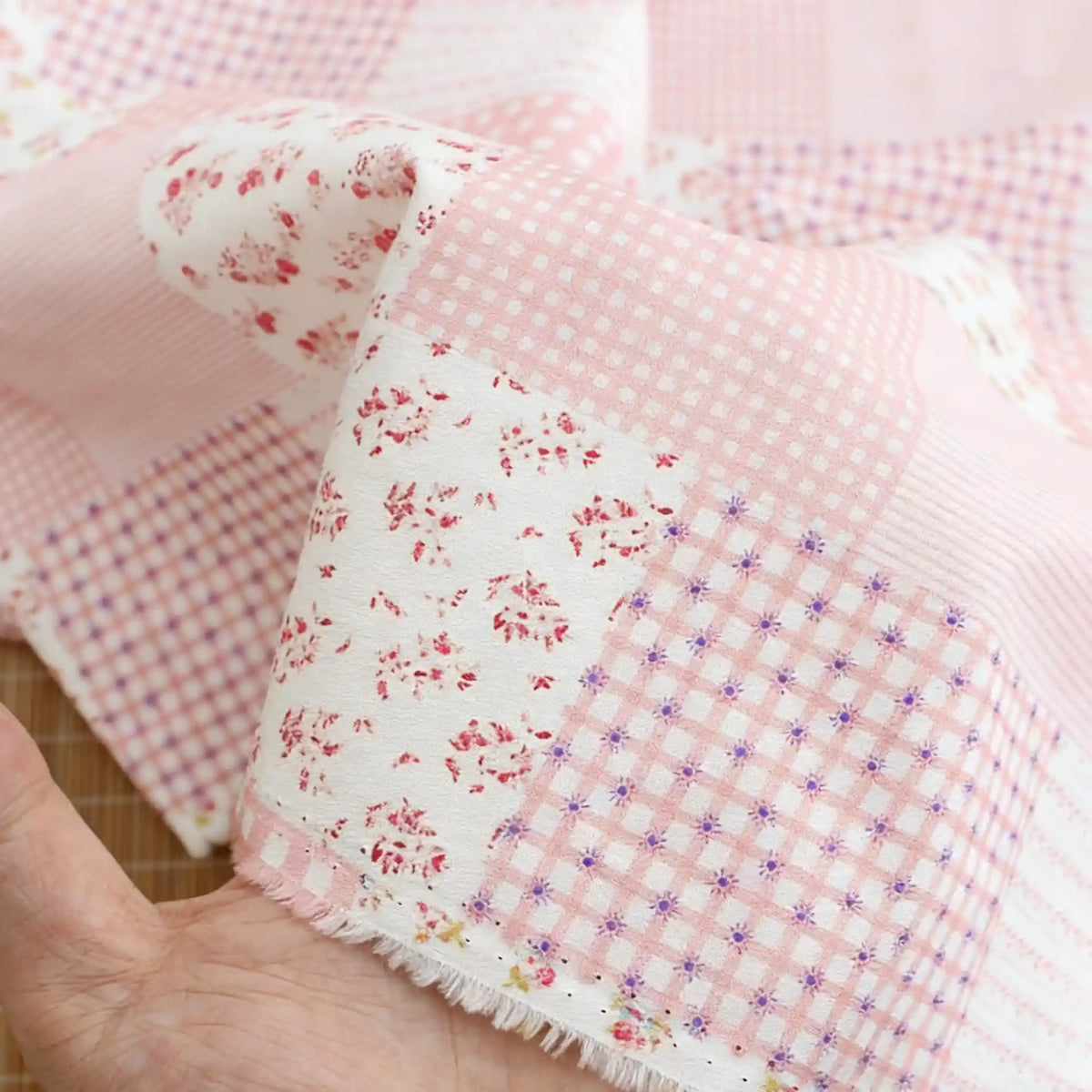 Une main tient le "Tissu popeline de coton style patchwork au mètre" de Longan Craft France, qui présente des motifs floraux et à carreaux pastel avec de charmants bords effilochés. Il s'agit d'une popeline de coton confortable, idéale pour les vêtements élégants.