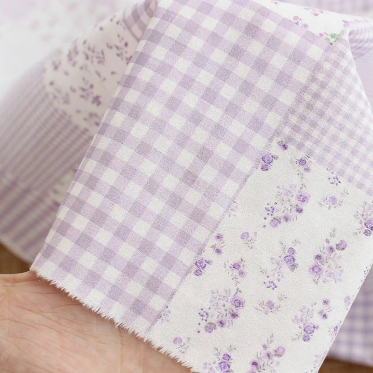 Une main tient deux morceaux de "Tissu popeline de coton style patchwork" de Longan Craft France : l'un à fleurs et carreaux pastel violets, l'autre blanc à petites fleurs violettes. Parfait pour vêtements stylés et confortables.