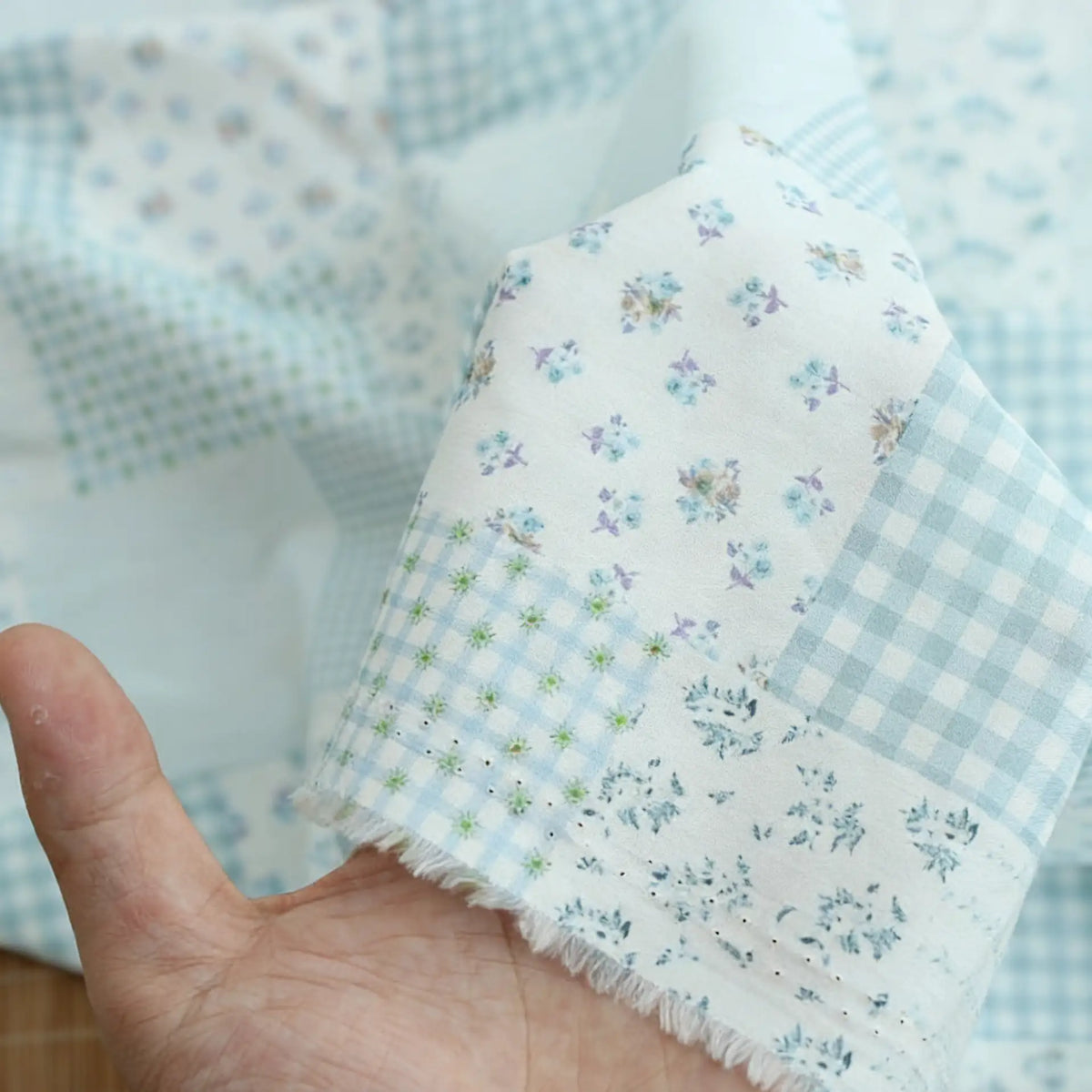 Une main présente le tissu popeline de coton patchwork au mètre de Longan Craft France, qui présente des motifs floraux et à carreaux pastel, y compris des motifs Vichy. Le bord du tissu est effiloché, ce qui met en valeur sa texture confortable pour des vêtements élégants.