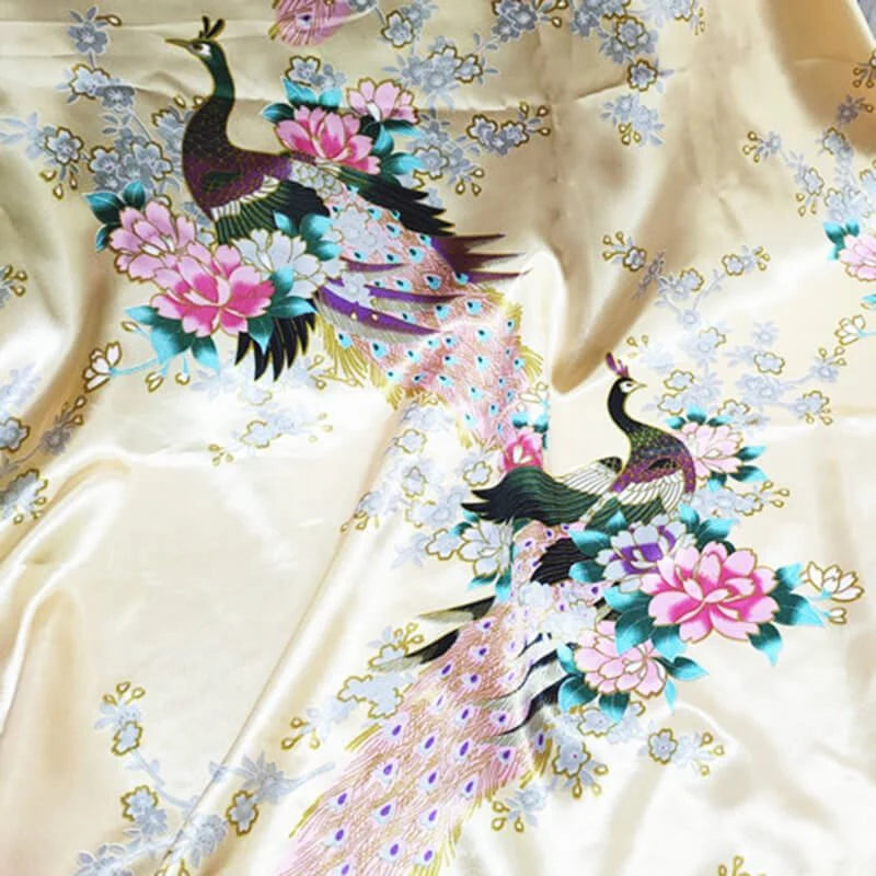 Tissu satin imprimé paon au mètre de Longan Craft France : tissu satiné crème orné de motifs paons colorés, grandes fleurs roses et touches bleues. Idéal pour une décoration intérieure élégante ou des accessoires raffinés.